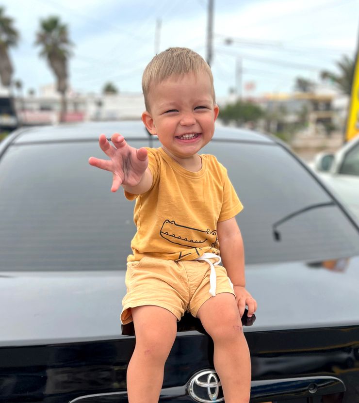 Amir, a toddler, smiling while sitting on the trunk of a car