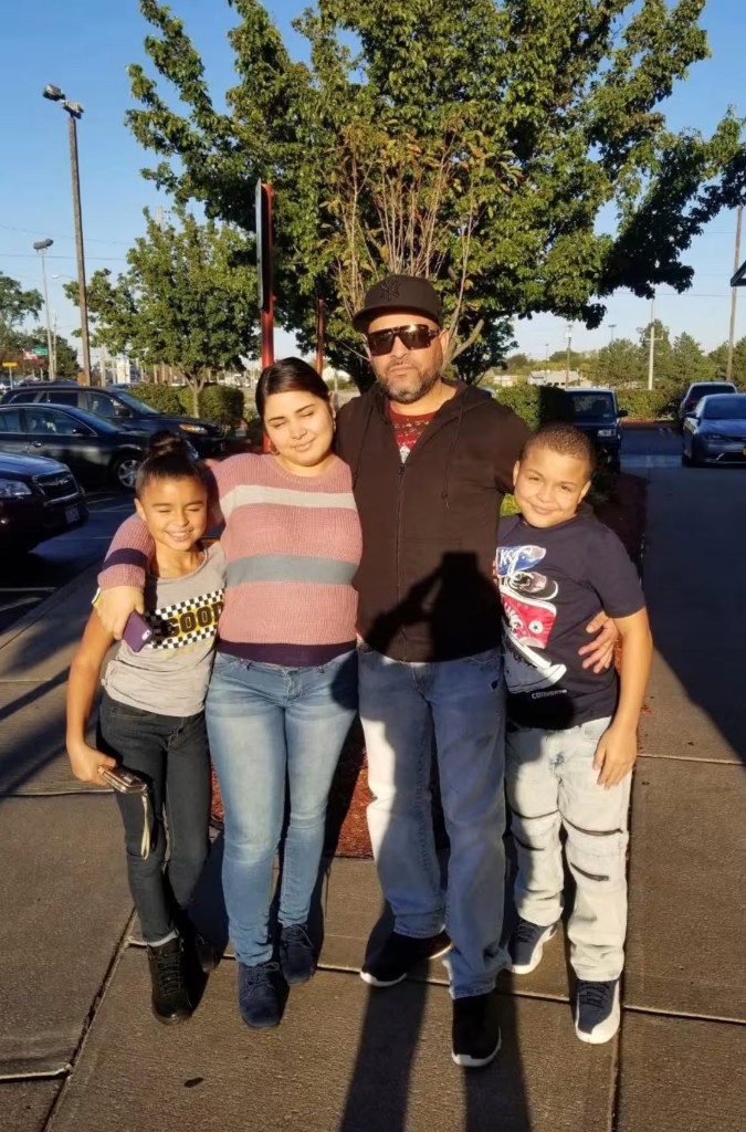 Geraldo Lunas Campos with his family outdoors, wearing a black hoodie and Yankees cap