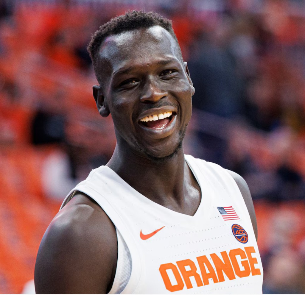 John Bol Ajak in a Syracuse Orange basketball jersey at an arena, smiling