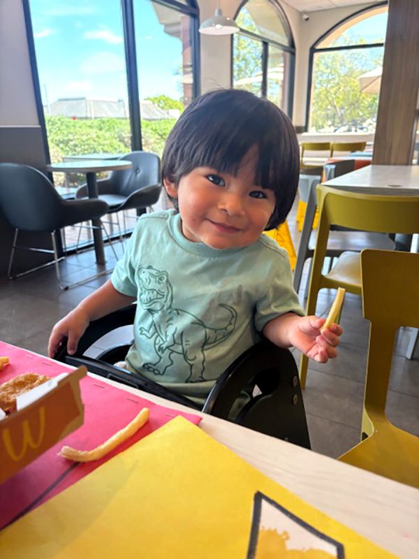 Kaleth, a toddler, smiling in a green dinosaur t-shirt at a restaurant table