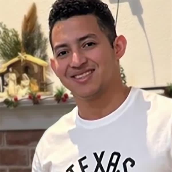 Neri Alvarado Borges smiling in a white t-shirt with TEXAS lettering, photographed indoors near a fireplace mantle
